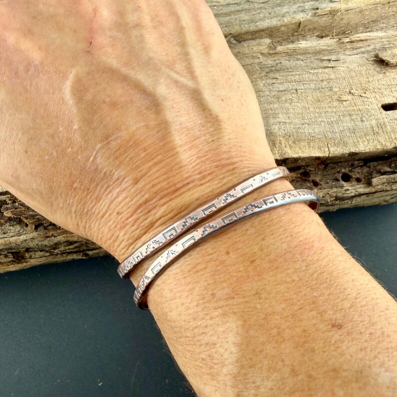 May include: Two thin, metallic cuff bracelets featuring a repeating geometric pattern engraved along the outer surface. The bracelets have a smooth, polished finish and a simple, open-ended design that wraps around the wrist.