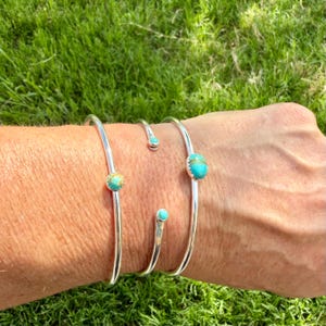 May include: Three silver-toned cuff bracelets with turquoise-colored stones. The bracelets are displayed on a person's arm against a backdrop of green grass. The turquoise stones are round and set in the center of the bracelets.