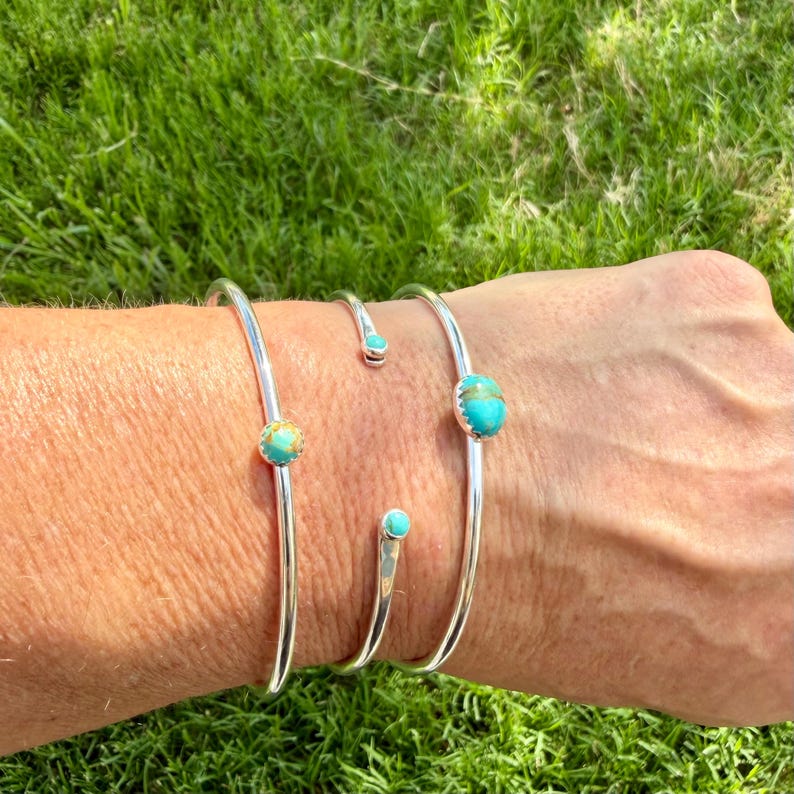 May include: Three silver-toned cuff bracelets with turquoise-colored stones. The bracelets are displayed on a person's arm against a backdrop of green grass. The turquoise stones are round and set in the center of the bracelets.