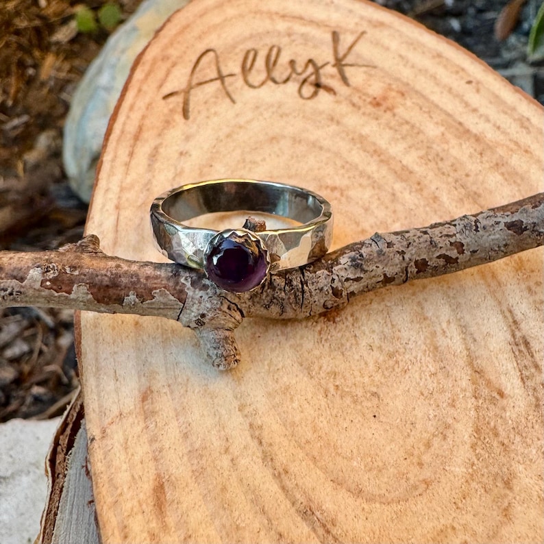 May include: A silver ring with a hammered texture and a round, deep purple gemstone. The ring is displayed on a small wooden branch, with the name "Allyk" written on the wooden surface in the background.