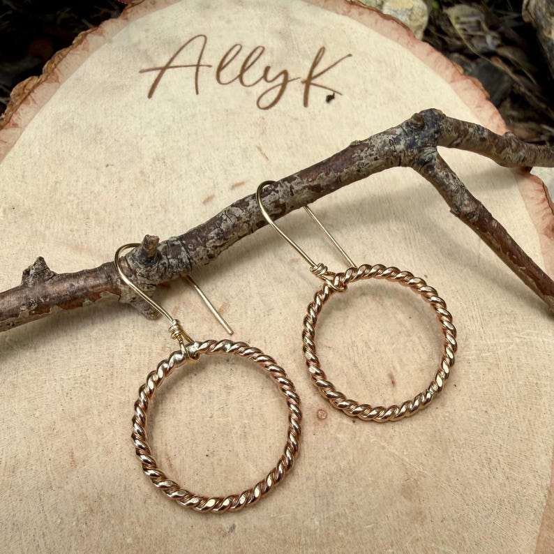 May include: A pair of gold-toned hoop earrings featuring a twisted metal texture. Each earring is suspended from a simple hook wire. The hoops are circular and have a polished finish, reflecting light across their twisted surface design.