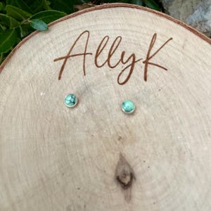 Pu&ograve; includere: Un paio di piccoli orecchini a bottone rotondi con montatura argentata e pietra color turchese. Gli orecchini sono esposti su una superficie di legno chiaro con il testo scritto a mano "Allyk".