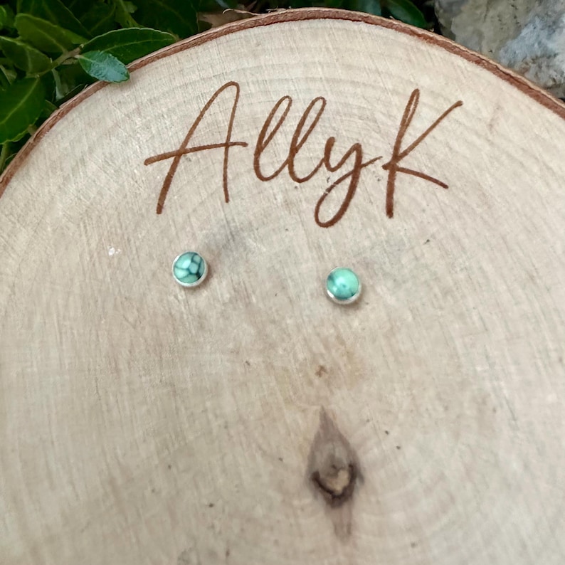 Pu&ograve; includere: Un paio di piccoli orecchini a bottone rotondi con montatura argentata e pietra color turchese. Gli orecchini sono esposti su una superficie di legno chiaro con il testo scritto a mano "Allyk".