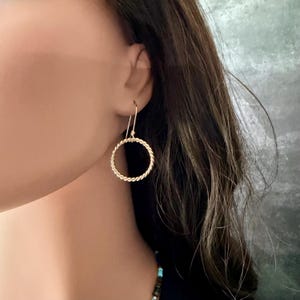 May include: A gold-toned earring featuring a circular hoop design with a twisted rope texture. The earring hangs from a simple hook wire, creating a dangling effect. The metal surface has a polished finish that reflects light, highlighting the intricate spiral pattern of the circular hoop component.