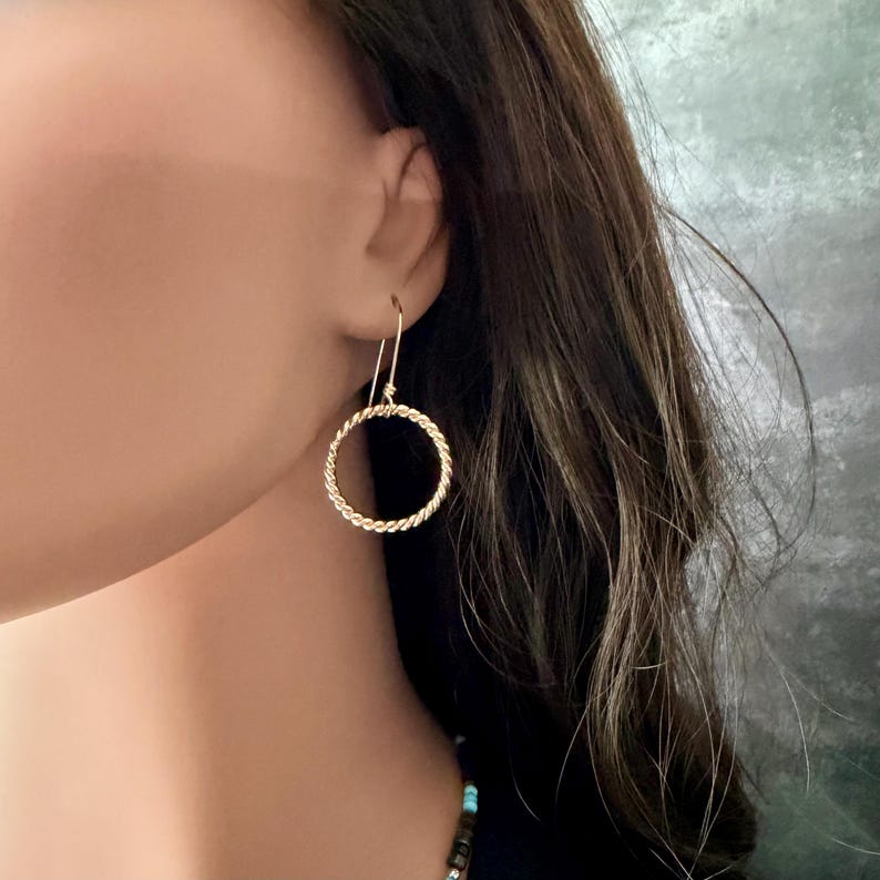 May include: A gold-toned earring featuring a circular hoop design with a twisted rope texture. The earring hangs from a simple hook wire, creating a dangling effect. The metal surface has a polished finish that reflects light, highlighting the intricate spiral pattern of the circular hoop component.