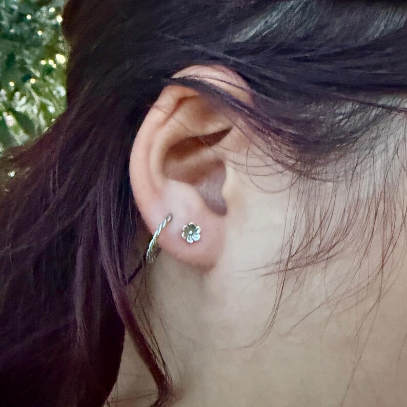 May include: A silver-toned earlobe piercing featuring a small, five-petaled flower stud earring and a thin, twisted silver-toned hoop earring positioned directly next to it on the lower edge of the earlobe.