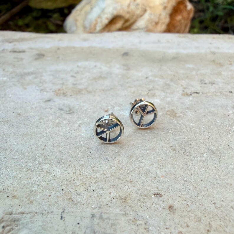 Puede incluir: Dos pendientes de bot&oacute;n plateados, cada uno con un dise&ntilde;o de s&iacute;mbolo de la paz. Los s&iacute;mbolos de la paz est&aacute;n delineados en un tono m&aacute;s oscuro, creando un contraste con el metal m&aacute;s claro. Los pendientes son redondos y tienen cierre de poste.