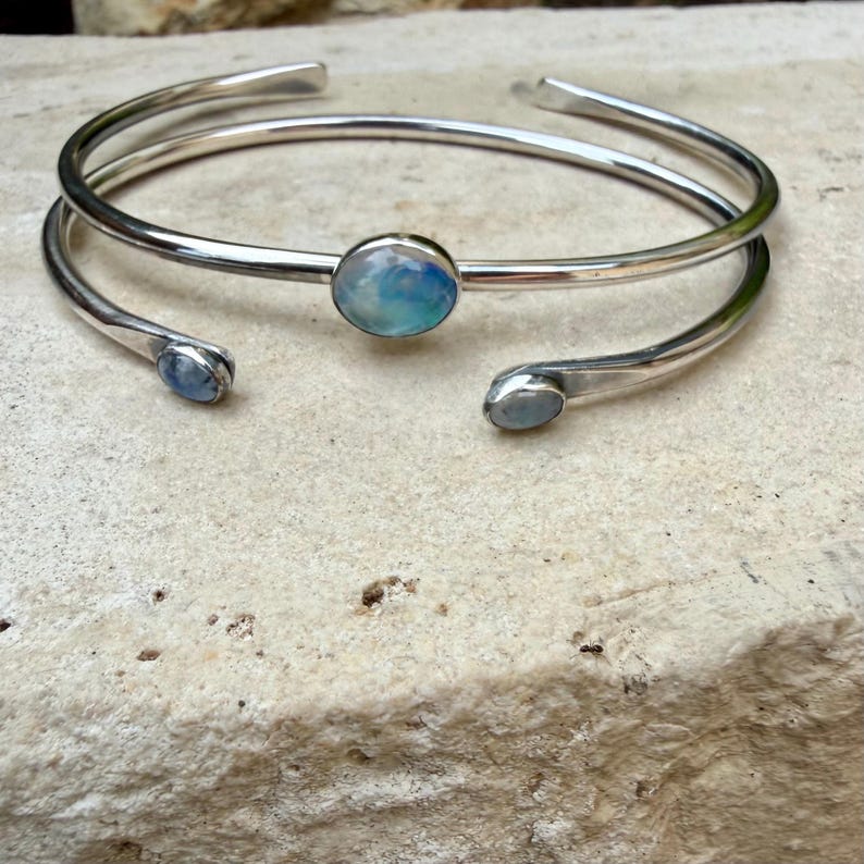 K&ouml;nnte beinhalten: Ein silbernes Armreif-Armband mit offenem Design. Das Armband hat einen gro&szlig;en, runden, irisierenden Opalstein in der Mitte und kleinere Opalsteine an den offenen Enden. Das Armband wird auf einer strukturierten, hellfarbigen Oberfl&auml;che pr&auml;sentiert.