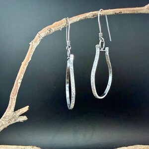 May include: A pair of silver teardrop earrings with a floral pattern, hanging from a branch against a dark background. The earrings have a simple, elegant design with a textured surface and a hook closure.