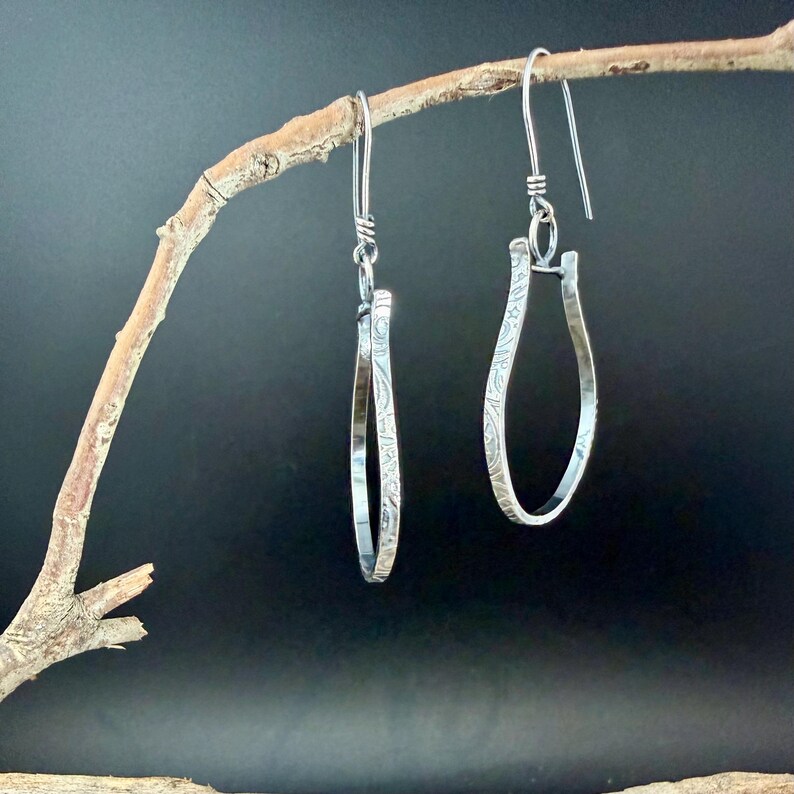 May include: A pair of silver teardrop earrings with a floral pattern, hanging from a branch against a dark background. The earrings have a simple, elegant design with a textured surface and a hook closure.