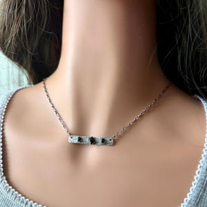 Pu&ograve; includere: Una collana a barra color argento con superficie testurizzata e incisa, caratterizzata da una pietra nera centrale a forma di diamante affiancata da due pietre nere rotonde pi&ugrave; piccole. Il pendente &egrave; appeso a una sottile catena a maglie argentata.