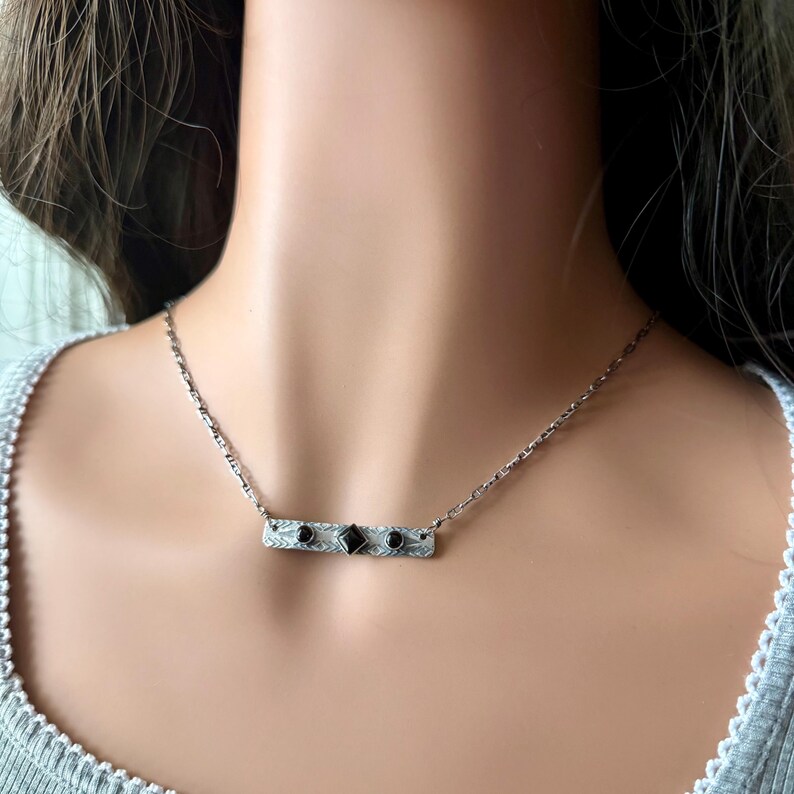 Pu&ograve; includere: Una collana a barra color argento con superficie testurizzata e incisa, caratterizzata da una pietra nera centrale a forma di diamante affiancata da due pietre nere rotonde pi&ugrave; piccole. Il pendente &egrave; appeso a una sottile catena a maglie argentata.