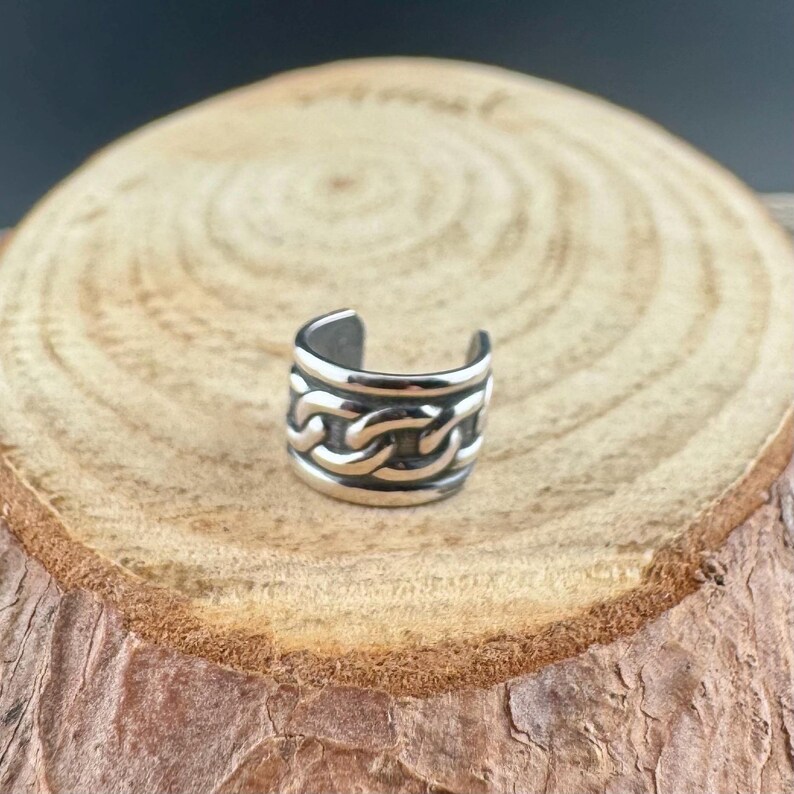 May include: A silver-colored ear cuff with a chain link design. The cuff is a cylinder shape with an opening at the top. The chain links are raised and create a textured surface. The cuff is sitting on a wooden surface.