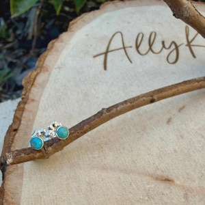 May include: A pair of silver stud earrings with turquoise stones. The earrings are displayed on a small branch, set against a wooden background. The word "Allyk" is written in cursive on the wood.