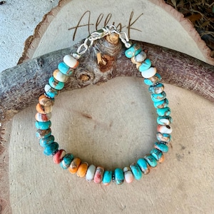 Pode incluir: Uma pulseira de contas com pedras redondas e planas em tons de turquesa, branco e laranja com veios castanhos. A pulseira &eacute; fixada com um fecho lagosta de metal prateado e inclui pequenas contas espa&ccedil;adoras de cor prateada.