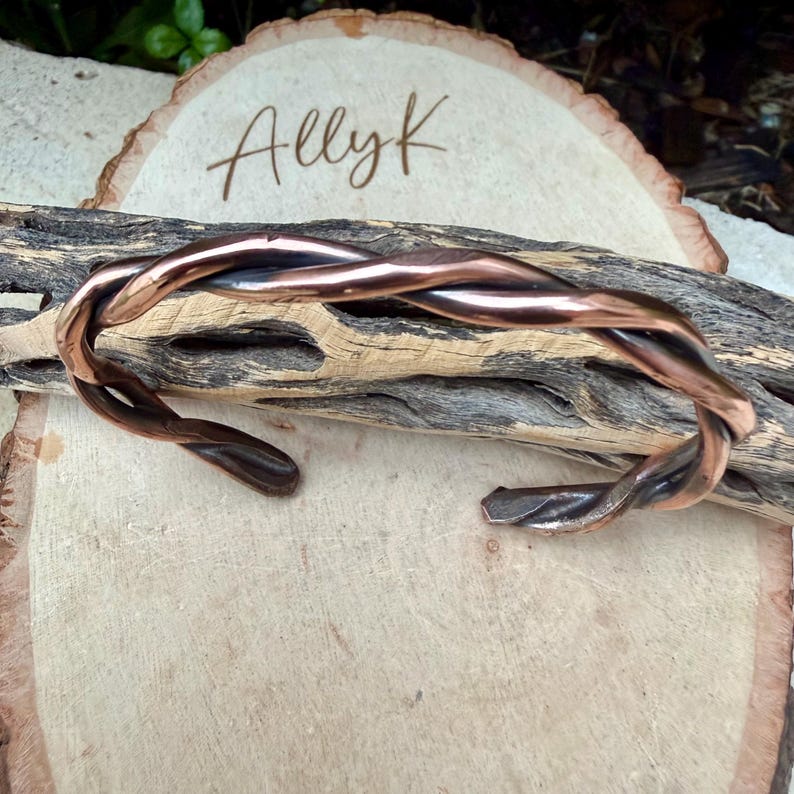 May include: A copper-colored cuff bracelet with a twisted design. The bracelet is open at the bottom and rests on a piece of wood. The name "Allyk" is written in cursive on a wooden slice in the background.