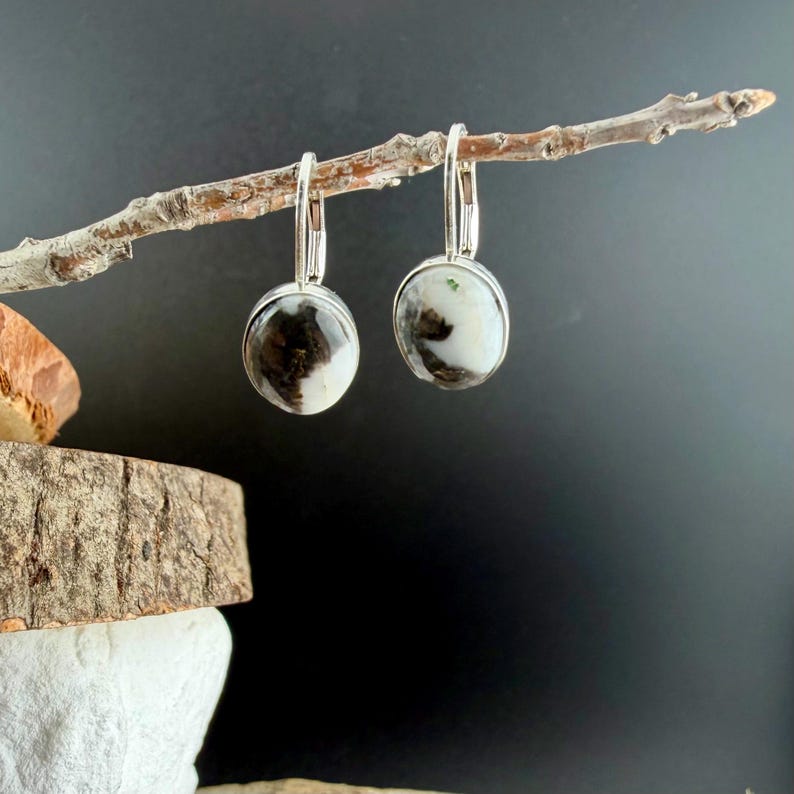 May include: A pair of silver leverback earrings with round, marbled stones. The stones feature a mix of white, black, and brown patterns. The earrings are displayed on a small, thin branch against a dark background.