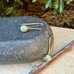 May include: A pair of gold-toned hook earrings featuring oval-shaped turquoise stones with brown matrix veining. The earrings have a simple, elongated wire design that curves to hold the stone setting securely at the base of each earring.