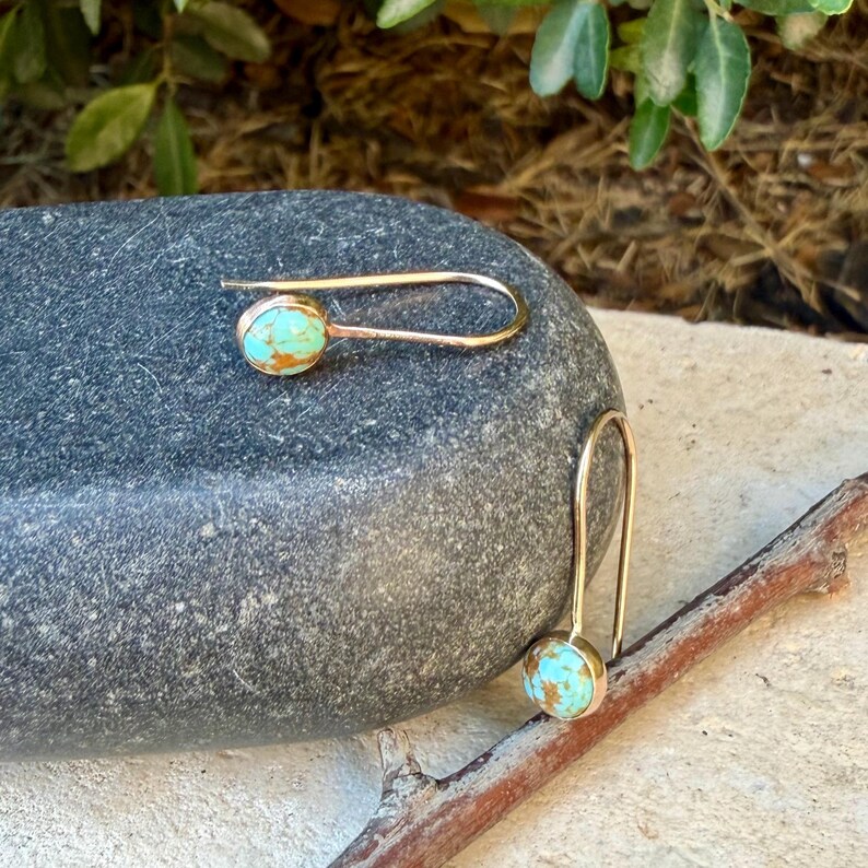 May include: A pair of gold-toned hook earrings featuring oval-shaped turquoise stones with brown matrix veining. The earrings have a simple, elongated wire design that curves to hold the stone setting securely at the base of each earring.