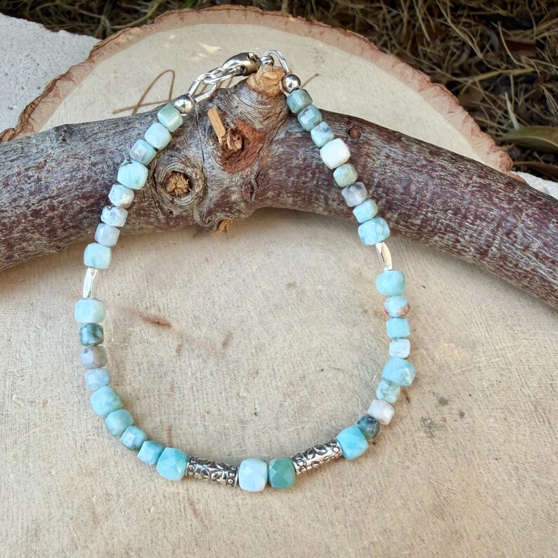 Puede incluir: Una pulsera compuesta por peque&ntilde;os cubos de piedra facetada en tonos azul claro y blanco, intercalados con dos piezas espaciadoras cil&iacute;ndricas de metal plateado. La pulsera se cierra con un broche de langosta de metal plateado.
