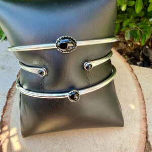 Op de afbeelding: Drie zilverkleurige manchetarmbanden met zwarte edelstenen. De bovenste armband heeft een ovale steen met een geschulpte rand. De andere twee hebben ronde stenen. De armbanden worden op een zwarte ondergrond gepresenteerd.