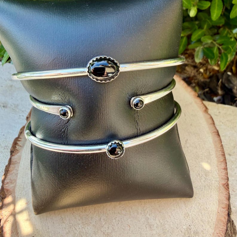 Op de afbeelding: Drie zilverkleurige manchetarmbanden met zwarte edelstenen. De bovenste armband heeft een ovale steen met een geschulpte rand. De andere twee hebben ronde stenen. De armbanden worden op een zwarte ondergrond gepresenteerd.