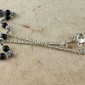 May include: A silver necklace with a chain and a clasp. The necklace features black beads and silver accents. The jewelry is displayed on a light brown wooden surface.