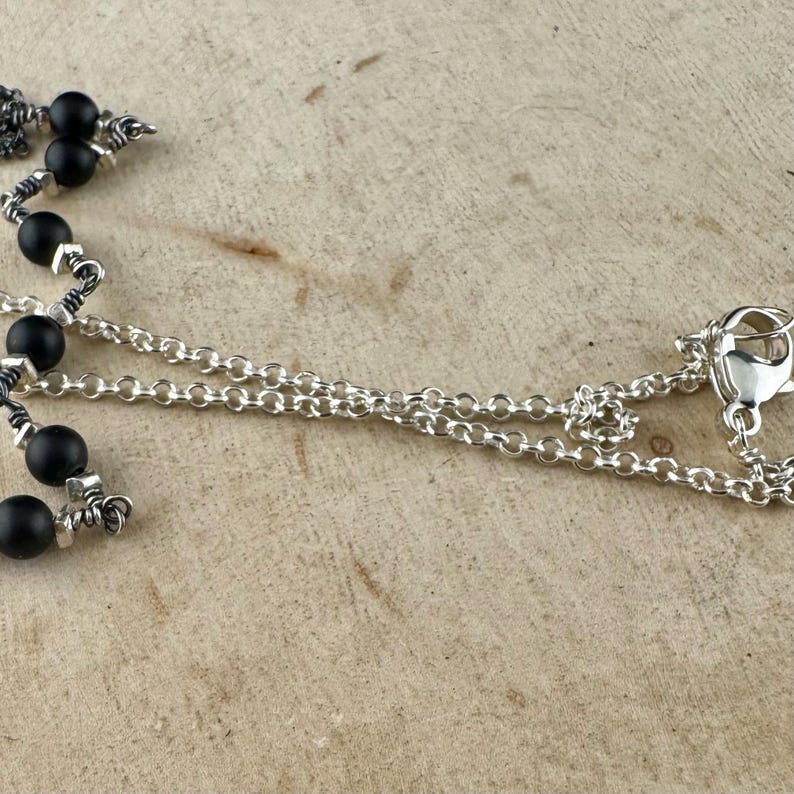 May include: A silver necklace with a chain and a clasp. The necklace features black beads and silver accents. The jewelry is displayed on a light brown wooden surface.
