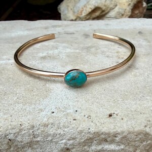 May include: A gold-colored cuff bracelet with an oval turquoise stone set in the center. The bracelet has a simple, open design. The turquoise stone has a mottled appearance with shades of blue and green.
