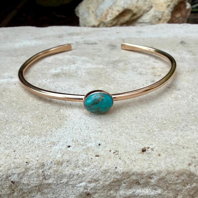 May include: A gold-colored cuff bracelet with an oval turquoise stone set in the center. The bracelet has a simple, open design. The turquoise stone has a mottled appearance with shades of blue and green.