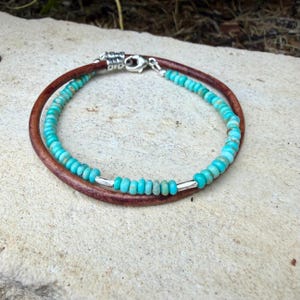 May include: A double-strand bracelet featuring one strand of smooth brown leather cord and one strand of small, round, turquoise-colored beads. The bracelet includes two small silver-toned metal spacer tubes and a silver-toned lobster claw clasp.