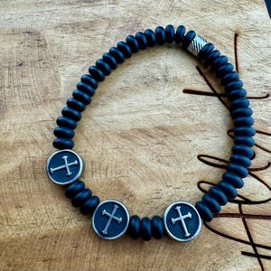 May include: A dark blue beaded bracelet with three silver-colored cross charms. The bracelet features a series of dark blue, rounded beads and a silver clasp. The cross charms are circular with a cross design.