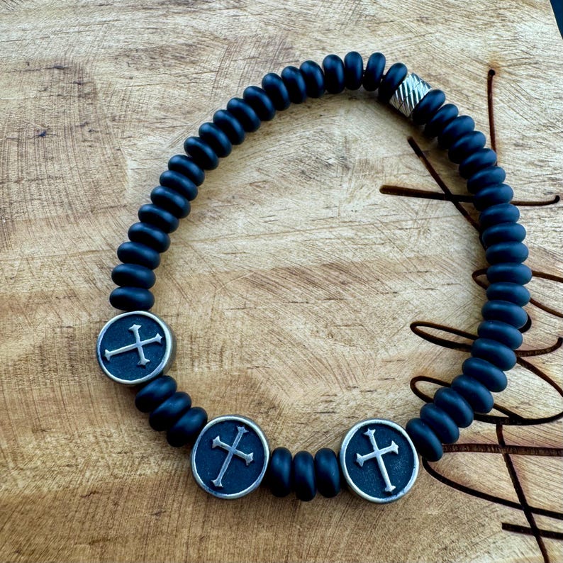 May include: A dark blue beaded bracelet with three silver-colored cross charms. The bracelet features a series of dark blue, rounded beads and a silver clasp. The cross charms are circular with a cross design.