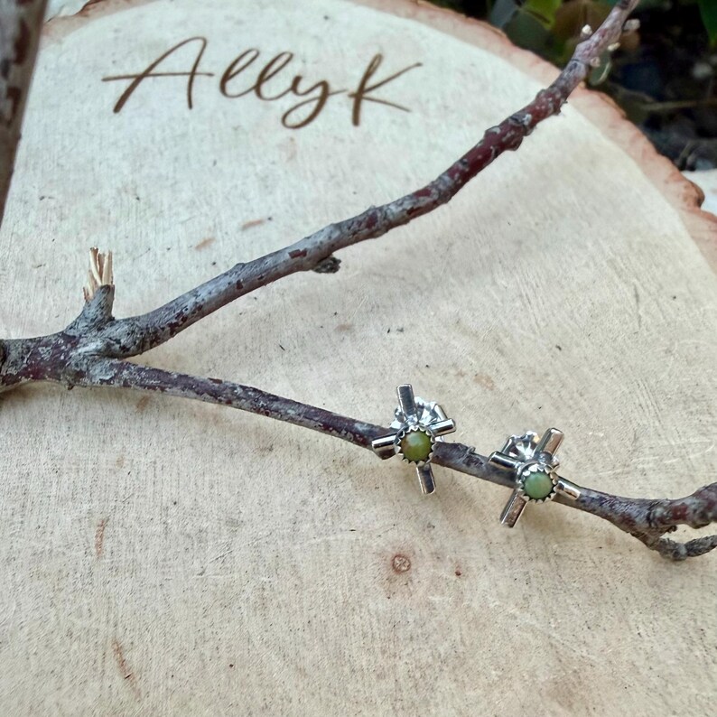 K&ouml;nnte beinhalten: Ein Paar silberfarbene Ohrstecker in Kreuzform mit einem kleinen, runden, hellgr&uuml;nen Stein in der Mitte jedes Kreuzes. Die Ohrringe werden auf einer Holzoberfl&auml;che mit dem handgeschriebenen Text "Allyk" pr&auml;sentiert.