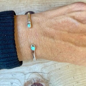 May include: A silver cuff bracelet featuring two open ends, each tipped with a small, oval-shaped turquoise stone. The metal band is smooth and polished, creating a simple, open-ended design that rests on the wrist.