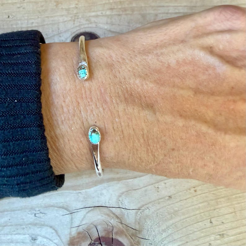 May include: A silver cuff bracelet featuring two open ends, each tipped with a small, oval-shaped turquoise stone. The metal band is smooth and polished, creating a simple, open-ended design that rests on the wrist.