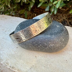 May include: A silver-toned metal cuff bracelet featuring an intricate, repeating embossed pattern of swirling lines, small stars, and circular motifs. The bracelet has a wide, open-ended band design with a polished finish that reflects light across its surface.