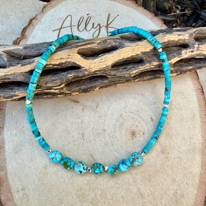 May include: Turquoise beaded necklace with silver accents. The necklace features a mix of small, square-cut turquoise beads and larger, round turquoise beads. The necklace is displayed on a wooden surface with the text "Allyk".