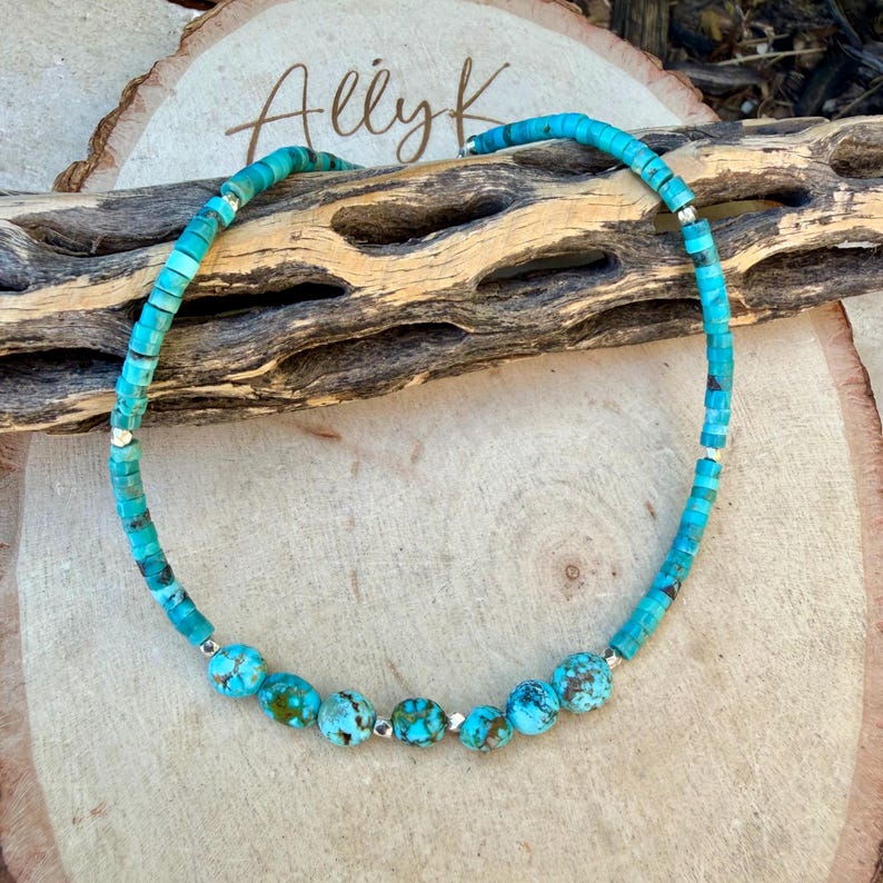 May include: Turquoise beaded necklace with silver accents. The necklace features a mix of small, square-cut turquoise beads and larger, round turquoise beads. The necklace is displayed on a wooden surface with the text "Allyk".