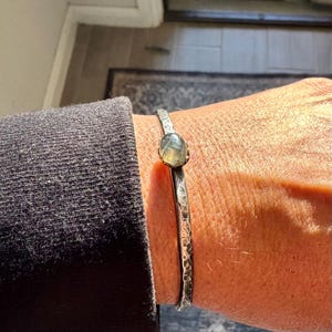 May include: A silver cuff bracelet with a hammered texture and a central, oval-shaped, light green gemstone. The bracelet is worn on a wrist, showcasing its design and craftsmanship.