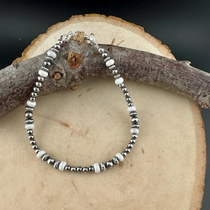 May include: A silver bracelet featuring alternating white and silver beads. The bracelet is composed of small, round silver beads and larger, oval-shaped white beads. The bracelet is displayed on a wooden surface.