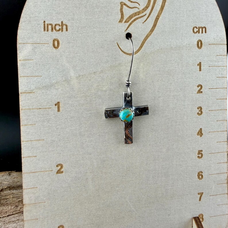 May include: A silver cross earring with a turquoise center stone. The cross is approximately 1 inch in length. The earring hangs from a silver wire. The cross has a textured surface.
