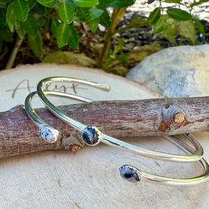 May include: Two silver-toned cuff bracelets with natural stone accents. One bracelet features three oval-shaped stones with black and white patterns. The other bracelet has a single round stone. The bracelets are displayed on a wooden branch.