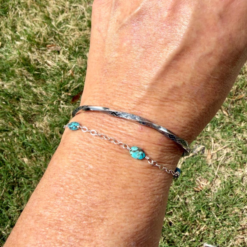 May include: Two bracelets on a wrist. The top bracelet is silver with a geometric pattern. The bottom bracelet is a silver chain with turquoise stones. The background is green grass.
