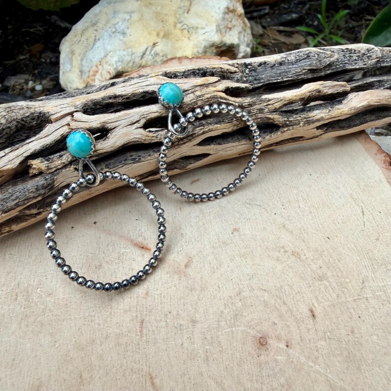 Puede incluir: Un par de pendientes de aro plateados con piedras turquesas. Los aros est&aacute;n hechos de peque&ntilde;as cuentas redondas. Los pendientes se exhiben sobre un trozo de madera flotante, mostrando una est&eacute;tica r&uacute;stica. Las piedras turquesas a&ntilde;aden un toque de color.