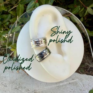 May include: Two silver ear cuffs displayed on a white ear-shaped stand. The top cuff features a shiny polished finish, while the bottom cuff has an oxidized polished finish. Both cuffs are engraved with a repeating leaf-like pattern.
