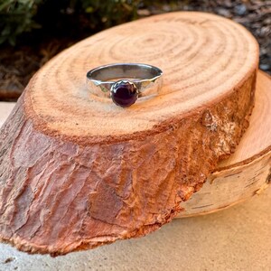 May include: A silver ring with a hammered band and a round, dark purple gemstone. The ring is set on a natural wood slice, showcasing the jewelry's design and colour.