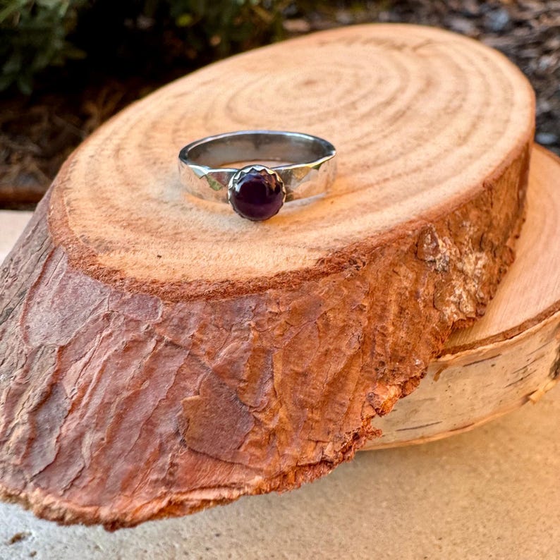 May include: A silver ring with a hammered band and a round, dark purple gemstone. The ring is set on a natural wood slice, showcasing the jewelry's design and colour.