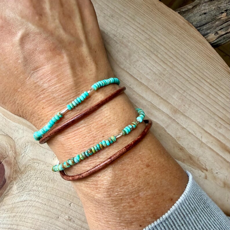 May include: A multi-strand bracelet set featuring two thin brown leather cords and two strands of small, round, turquoise-colored beads. The turquoise beads are interspersed with tiny silver-toned metal spacers, creating a layered look.
