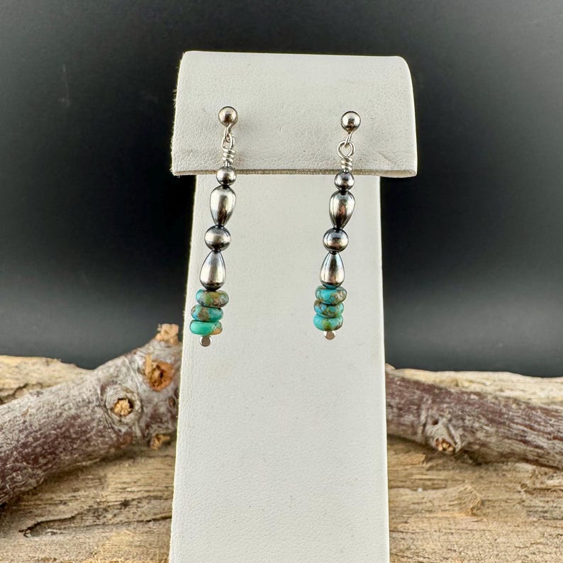 May include: Silver dangle earrings with a series of silver beads and turquoise stones. The earrings have a post backing and are displayed on a white stand against a dark background.